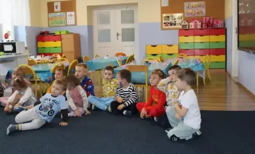 Dzieci w grupie siedzą na podłodze na ciemnej wykładzinie w sali zabaw.
