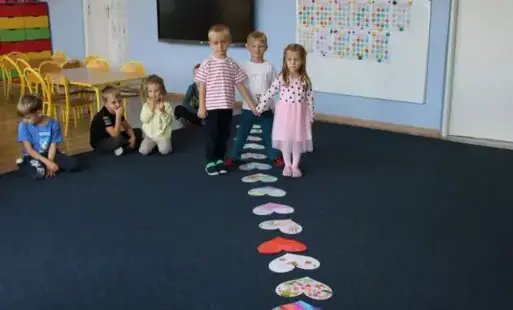 Chłopiec i dziewczynka trzymają się za ręce i idą nad rozłożonymi kartkami w kształcie serc na wykładzinie, pozostałe dzieci siedzą obok.