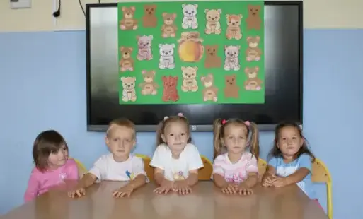 Dzieci siedzą przy stole, a za plecami na ścianie na tablicy przypięte są misie pokolorowane kredkami i wycięte z kartek